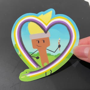 Pode incluir: Um personagem de pincel com um corpo de pincel amarelo e marrom, segurando um pincel menor, está rodeado por um contorno em forma de coração da bandeira do orgulho não binário. O fundo é um campo verde com um céu azul.