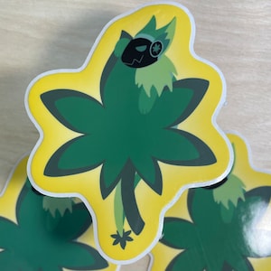 May include: A yellow sticker with a green four-leaf clover design. A cartoon character with a black mask and green fur is inside the clover.