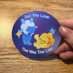 May include: Round sticker with a blue background featuring two cartoon flowers. One is blue with white petals, the other is yellow. Text reads "But You Love The Way She Lies".