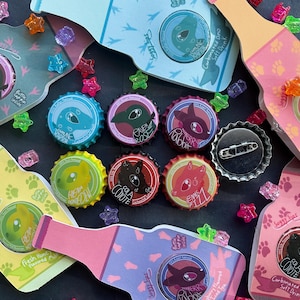 Könnte beinhalten: Eine Sammlung bunter Flaschenverschlüsse mit verschiedenen Dinosaurier-Designs und Namen wie "Hiena Hovenden", "Raven Rose", "Wobbery Rabbit", "Ennes Fizz", "Cola Coyote" und "Marine Raptor". Die Flaschenverschlüsse sind auf einer schwarzen Oberfläche angeordnet, mit einigen bunten Plastikperlen, die um sie herum verstreut sind.