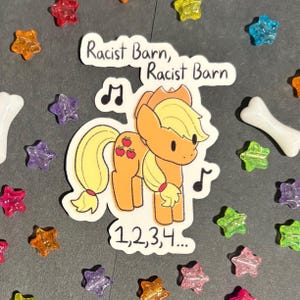 May include: A sticker featuring a cartoon pony with a blonde mane and tail, red apple markings, and musical notes. The text "Racist Barn" and "1,2,3,4..." are also present. The sticker is surrounded by colorful star-shaped beads and a bone-shaped bead.