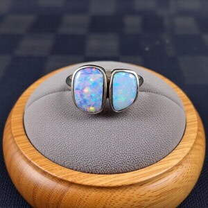 May include: A silver ring featuring two large, iridescent opal stones. The opals display a range of colors, including blue, purple, and green. The ring is set on a gray velvet surface, displayed on a wooden stand.