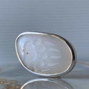 May include: A white oval-shaped ring with a silver band. The ring features a white bear paw print design.