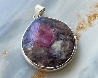 May include: A silver pendant featuring a large, faceted, purple and pink gemstone with a white center. The pendant is on a white marble surface.