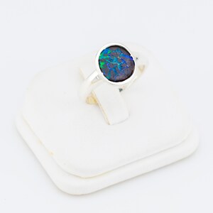 May include: A silver ring featuring a dark opal stone with blue and green iridescent patterns. The oval-shaped stone is set in a bezel setting, and the ring is displayed on a white surface.