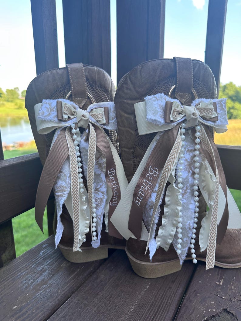 May include: Two brown cowboy boots decorated with cream-coloured bows, floral patterns, and pearl strands. Brown ribbons with the name "Bethany" are attached. The boots are set against a natural outdoor backdrop.