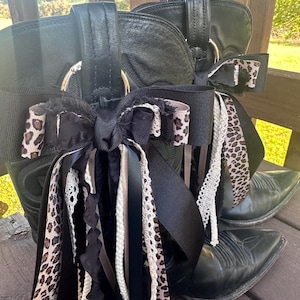 Puede incluir: Un par de botas de vaquero de cuero negro adornadas con lazos y cintas decorativas. Los lazos presentan un estampado de leopardo, y las cintas incluyen detalles en negro, blanco y beige. Las botas tienen puntas puntiagudas.
