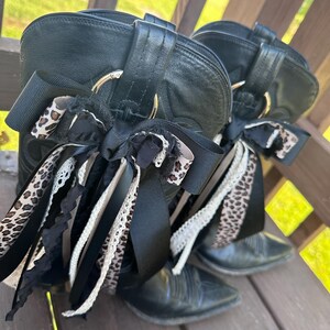 Puede incluir: Un par de botas de vaquero de cuero negro con detalles de cintas decorativas. Las cintas presentan estampado de leopardo, detalles de encaje negro y blanco. Las botas est&aacute;n sobre una superficie de madera.