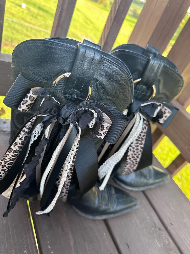 Puede incluir: Un par de botas de vaquero de cuero negro con detalles de cintas decorativas. Las cintas presentan estampado de leopardo, detalles de encaje negro y blanco. Las botas est&aacute;n sobre una superficie de madera.