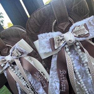 May include: Brown suede cowboy boots with decorative bows. The bows have cream floral fabric, brown ribbons, and beaded details. The name "Bethany" is embroidered on the ribbons.