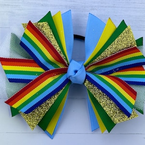 May include: A rainbow hair bow with blue, green, yellow, and gold glitter ribbon. The bow is tied with a light blue ribbon and has a white tulle accent.