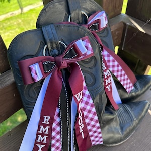 May include: A pair of black leather cowboy boots adorned with burgundy and white ribbons. The ribbons feature the names "Emmy" and "Woopi" in white lettering, along with a gingham pattern. The boots are resting on a wooden surface.