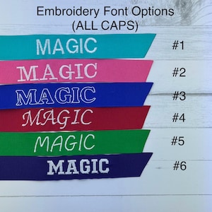 May include: Image of six coloured ribbons, each with the word "MAGIC" embroidered in white. The ribbons are teal, pink, blue, red, green, and purple. The text "Embroidery Font Options (ALL CAPS)" is at the top, with numbers 1-6 next to each ribbon.