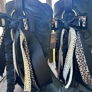 Puede incluir: Un par de botas de vaquero de cuero negro adornadas con lazos decorativos. Los lazos presentan cintas de sat&eacute;n negro, estampado de leopardo y encaje color crema. Anillos met&aacute;licos dorados est&aacute;n unidos a la parte superior de las botas.