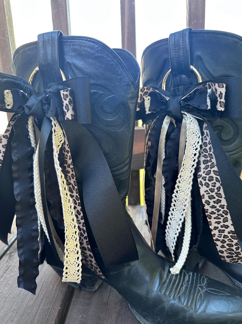 Puede incluir: Un par de botas de vaquero de cuero negro adornadas con lazos decorativos. Los lazos presentan cintas de sat&eacute;n negro, estampado de leopardo y encaje color crema. Anillos met&aacute;licos dorados est&aacute;n unidos a la parte superior de las botas.