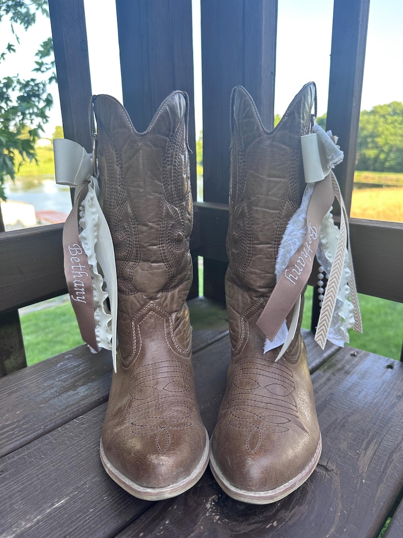 May include: A pair of brown cowboy boots with decorative stitching and a cream-coloured sole. Each boot has a cream-coloured bow and a ribbon with the name "Bethany" printed on it. The boots are set on a wooden surface.