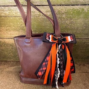 May include: A brown leather tote bag with long handles. A decorative bow with orange, black, and white ribbons and lace is attached to the bag's handle. The bow features a plaid pattern and adds a touch of flair to the bag.