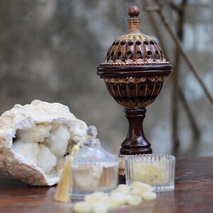 May include: A wooden incense burner with a decorative gold design, a clear glass jar with a gold tassel, a white geode, and a small glass dish filled with yellow resin.