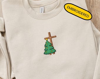 May include: A beige sweatshirt with an embroidered design featuring a brown cross, a green Christmas tree, and a yellow star. The word "EMBROIDERED" is printed on a yellow tag. The sweatshirt is folded.