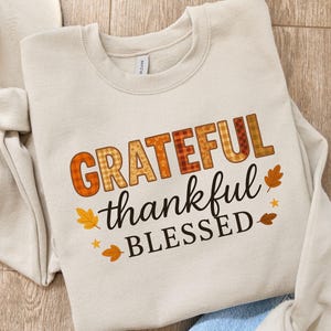 Peut inclure: Sweat-shirt beige avec l'inscription "GRATEFUL thankful BLESSED" en orange et noir. Le mot "GRATEFUL" présente un motif à carreaux. Des feuilles d'automne et des étoiles entourent le texte. Le sweat-shirt est plié sur une surface en bois.