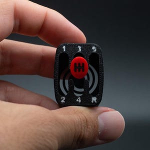 May include: A black and red gear shift knob with the numbers 1, 2, 3, 4, 5, and R printed in white. The knob has a red circular button in the centre and is held in a hand.