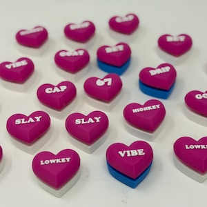 May include: A collection of pink heart-shaped objects with white text, including words like "VIBE", "SLAY", and "GOAT". Some hearts have a blue base, while others have a white base. The objects are arranged on a white surface.
