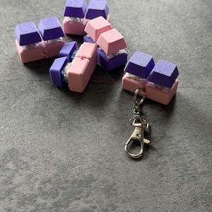 May include: A collection of colorful keycap-shaped accessories in pink and purple hues. The keycaps are attached to a silver-colored keychain clasp. The items are scattered on a gray surface.