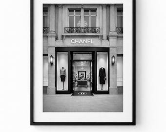 May include: Black and white photograph of a Chanel store exterior. The store entrance is framed by large windows displaying mannequins. The word "CHANEL" is displayed above the entrance. The building has a classic architectural design.