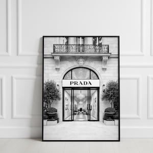 以下が含まれることがあります： プラダの店舗外観の白黒フレームプリント。建物は、入り口の上にプラダのロゴが付いたアーチ型の入り口が特徴です。入り口の両側には木と装飾的なプランターがあります。装飾的な手すりのあるバルコニーが上に表示されます。
