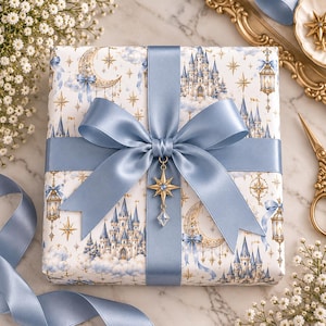 May include: A square gift box wrapped in white paper featuring a pattern of blue castles, golden crescent moons, stars, and lanterns. The box is tied with a wide, dusty blue satin ribbon and a large bow, accented by a gold star-shaped charm.
