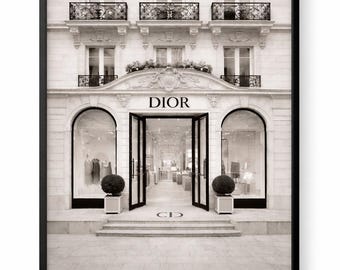 May include: Monochrome framed print of the Dior boutique facade. The building has arched windows, balconies, and the Dior logo above the entrance. Two topiary balls are positioned either side of the entrance. A classic, elegant image representing the brand.