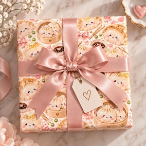 May include: A square gift box wrapped in paper featuring a repeating pattern of cute, smiling dumplings in bamboo steamers, pink bows, and small hearts. The box is tied with a wide, dusty pink satin ribbon and a small white gift tag with a heart.
