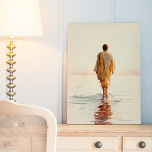 May include: A canvas print showing a figure walking in water, seen from behind. The person wears a light-coloured robe, with the water reflecting the figure and the sky. The artwork is displayed on a light-coloured wooden surface.