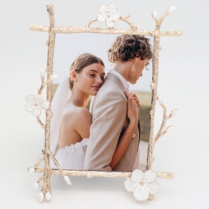 May include: A gold-colored metal picture frame with a floral branch design. The frame holds a photograph of a couple embracing. The frame has white flower accents and a rectangular shape.