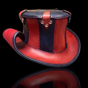 May include: A handcrafted top hat in red and navy blue leather. The hat features a curved brim and is stitched with contrasting thread. Silver-toned rivets secure the leather strap.