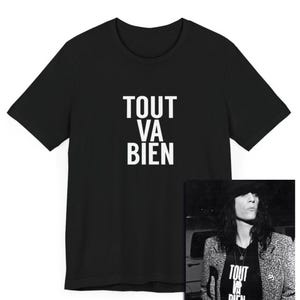 May include: Black t-shirt with white text that reads "Tout Va Bien". The shirt is shown on a model wearing a black jacket and a t-shirt with the same text.