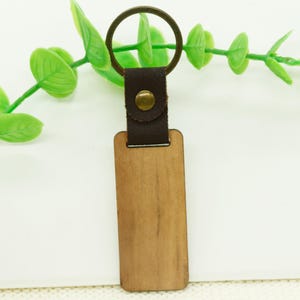 以下が含まれることがあります： 茶色の革ひもに金属製のリングが付いた木製キーホルダー。キーホルダーは空白で、パーソナライズできます。