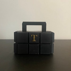 Puede incluir: Un objeto negro, geométrico, en forma de caja con un asa. El objeto está construido a partir de múltiples bloques cuadrados. Una letra "T" de color dorado está centrada en la parte delantera. El objeto está sobre una superficie oscura.