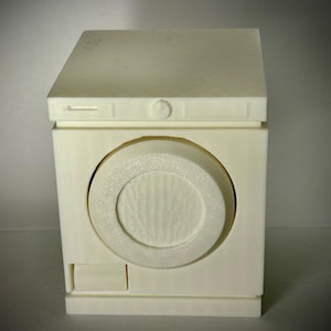 May include: A small, off-white model of a washing machine. The appliance has a rectangular shape with a circular door and a small rectangular compartment below. The top has a slightly angled design.