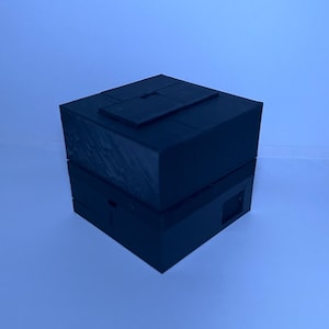 May include: A black, geometric box with a sliding top and a rectangular opening on the side. The box is made of a solid, dark material and sits against a light blue background. The design suggests a storage container or a puzzle box.