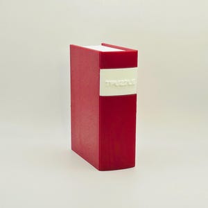 Puede incluir: Un objeto rojo con forma de libro que tiene una etiqueta blanca en el lomo con el texto en relieve TPUZZLE. El artículo presenta una estructura rectangular vertical con un acabado liso de color sólido y un borde superior blanco.