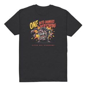 Puede incluir: Camiseta negra con un monstruo de dibujos animados con dientes afilados y el texto "ONE BITE CHANGES EVERYTHING" en amarillo y rojo. La frase "ALPHA GAL SYNDROME" está impresa debajo.