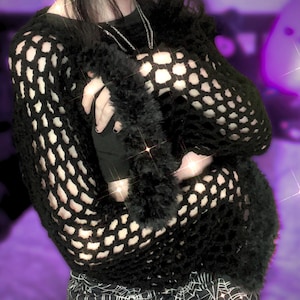 May include: Black open-knit sweater with a fuzzy trim. The sweater has a unique, holey design. The sleeves are long and the overall style is edgy and alternative. The sweater is paired with a black top underneath and a skirt with a spiderweb pattern.