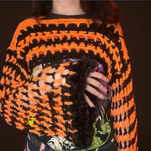 May include: An orange and black crochet top with a unique open weave design. The sleeves are trimmed with black faux fur. A neon green safety pin is attached to the top. The top is worn over a graphic t-shirt.