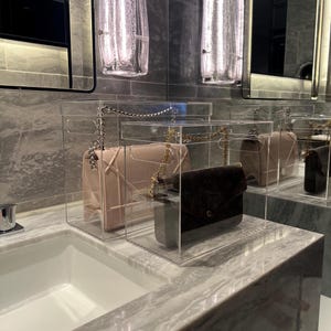 May include: Three clear acrylic boxes with lids, each containing a designer handbag. The handbags are a light pink, a dark brown, and a black.