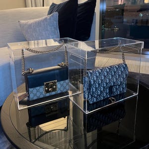 Puede incluir: Dos bolsos de diseñador se muestran en cajas de acrílico transparentes. El bolso de la izquierda es negro con un patrón marrón y blanco. El bolso de la derecha es azul con un patrón blanco.