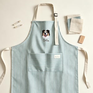 May include: A light blue and white striped apron featuring an embroidered Australian Shepherd dog face above the name Bella. The apron includes a front pocket, cream-coloured neck and waist ties, an adjustable neck buckle, and two silver safety pins.