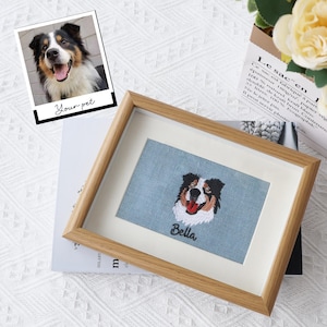 May include: A rectangular wooden frame containing a light blue fabric panel with a detailed embroidery of a black, white, and brown dog with its tongue out. Below the dog, the name Bella is embroidered in black script on the blue fabric.