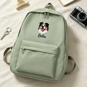 以下が含まれることがあります： セージグリーンのバックパック。黒、白、茶色の犬の刺繍が施されており、その下に黒の筆記体でBellaという名前が刺繍されています。バックパックにはフロントジッパーポケットとシルバーの金属製ジッパーが付いています。
