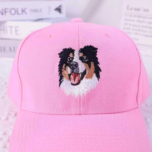 May include: A pink baseball cap featuring an embroidered image of a dog's face. The dog has black, brown, and white fur, with its mouth open. The cap is made of a soft fabric and has a curved brim.